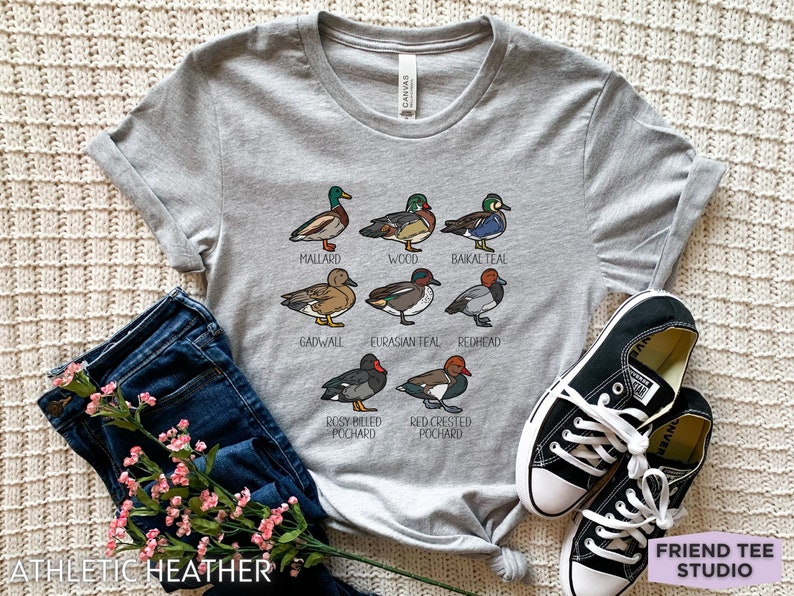 Peut inclure: Un t-shirt gris chin&eacute; avec une impression graphique de huit esp&egrave;ces de canards diff&eacute;rentes, accompagn&eacute;es de leurs noms respectifs : Mallard, Wood, Baikal Teal, Gadwall, Eurasian Teal, Redhead, Rosy Billed Pochard et Red Crested Pochard.