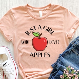 May include: A peach-colored t-shirt with the text "JUST A GIRL WHO LOVES APPLES" in black. A red apple with a green leaf is in the center. White sneakers with red trim and blue jeans are visible.