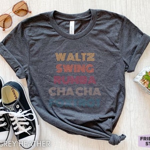 May include: Dark grey heather t-shirt with the words "Waltz, Swing, Rumba, Cha Cha" in retro-style lettering. The shirt is paired with black sneakers and sunglasses. The text "Friend Tee Studio" is also visible.