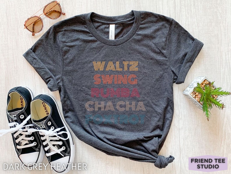May include: Dark grey heather t-shirt with the words "Waltz, Swing, Rumba, Cha Cha" in retro-style lettering. The shirt is paired with black sneakers and sunglasses. The text "Friend Tee Studio" is also visible.
