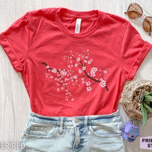 Puede incluir: Una camiseta roja jaspeada con un gr&aacute;fico de una rama de flor de cerezo. La camiseta est&aacute; hecha de un material suave y c&oacute;modo y tiene un corte holgado.