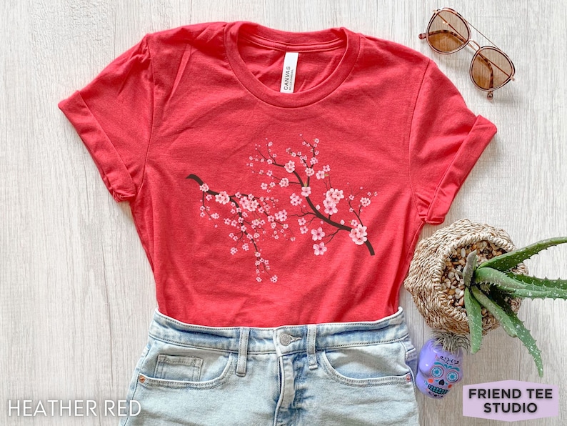 Puede incluir: Una camiseta roja jaspeada con un gr&aacute;fico de una rama de flor de cerezo. La camiseta est&aacute; hecha de un material suave y c&oacute;modo y tiene un corte holgado.