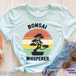 May include: A light blue t-shirt with a vintage-style graphic of a bonsai tree and the text "Bonsai Whisperer".
