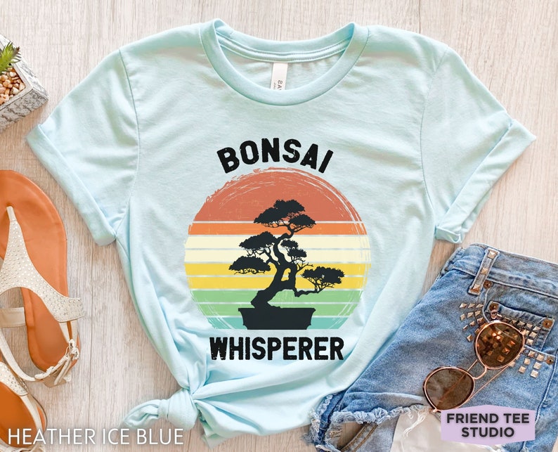 May include: A light blue t-shirt with a vintage-style graphic of a bonsai tree and the text "Bonsai Whisperer".