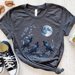 Peut inclure: T-shirt gris foncé avec un motif de trois corbeaux noirs perchés sur une branche avec des feuilles bleues, une pleine lune et un ciel bleu. Le t-shirt est en matière souple et a un col rond.