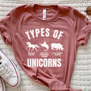 May include: A pink t-shirt with the text "Types of Unicorns" in white. The shirt features illustrations of a unicorn, a swordfish, and a rhinoceros, each labeled with a different type of unicorn: Fairytale Unicorn, High Speed Submersible Unicorn, and Heavy Assault Unicorn.