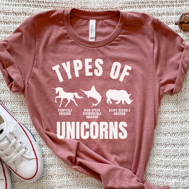 May include: A pink t-shirt with the text "Types of Unicorns" in white. The shirt features illustrations of a unicorn, a swordfish, and a rhinoceros, each labeled with a different type of unicorn: Fairytale Unicorn, High Speed Submersible Unicorn, and Heavy Assault Unicorn.