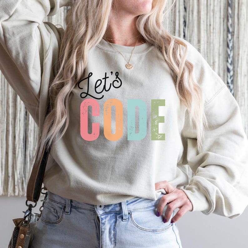 K&ouml;nnte beinhalten: Ein hellbeige Sweatshirt mit dem Text "Let's CODE" in schwarzen und bunten Buchstaben. Das Wort "CODE" ist in Rosa, Orange, Blau und Gr&uuml;n gehalten. Das Sweatshirt wird mit einer blauen Jeans kombiniert.