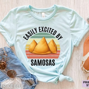 Puede incluir: Una camiseta azul claro con un gráfico de tres samosas doradas centradas sobre un círculo de rayas estilo retro. El texto "EASILY EXCITED BY SAMOSAS" está impreso en letras mayúsculas negras, arqueándose sobre y bajo la ilustración.