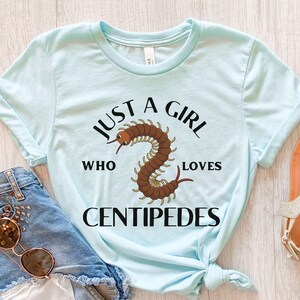 May include: Light blue t-shirt with the text "JUST A GIRL WHO LOVES CENTIPEDES" in black. The shirt features a brown centipede illustration. The shirt is made of a soft material and has short sleeves.