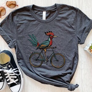 K&ouml;nnte beinhalten: Dunkelgraues T-Shirt mit einer farbenfrohen Illustration eines Vogels auf einem Fahrrad. Der Vogel hat rote, t&uuml;rkisfarbene und gelbe Details. Das Fahrrad ist schwarz mit goldfarbenen R&auml;dern. Das Shirt ist aus weichem Material.
