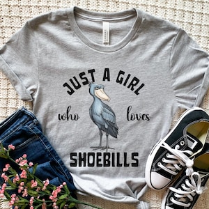 Puede incluir: Camiseta gris con una ilustraci&oacute;n de dibujos animados de un zoc&oacute; y el texto "Just a girl who loves shoebills".