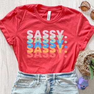 May include: A red t-shirt with the word "SASSY" stacked vertically in colorful letters. The shirt is paired with denim shorts. Sunglasses and a decorative basket are visible in the background.