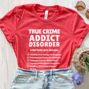 May include: A red t-shirt with the text "TRUE CRIME ADDICT DISORDER" in white. The shirt lists symptoms including "Thinking Every Stranger Is Suspicious" and "Googling 'Untraceable Poisons' For Fun". The shirt is paired with denim shorts.