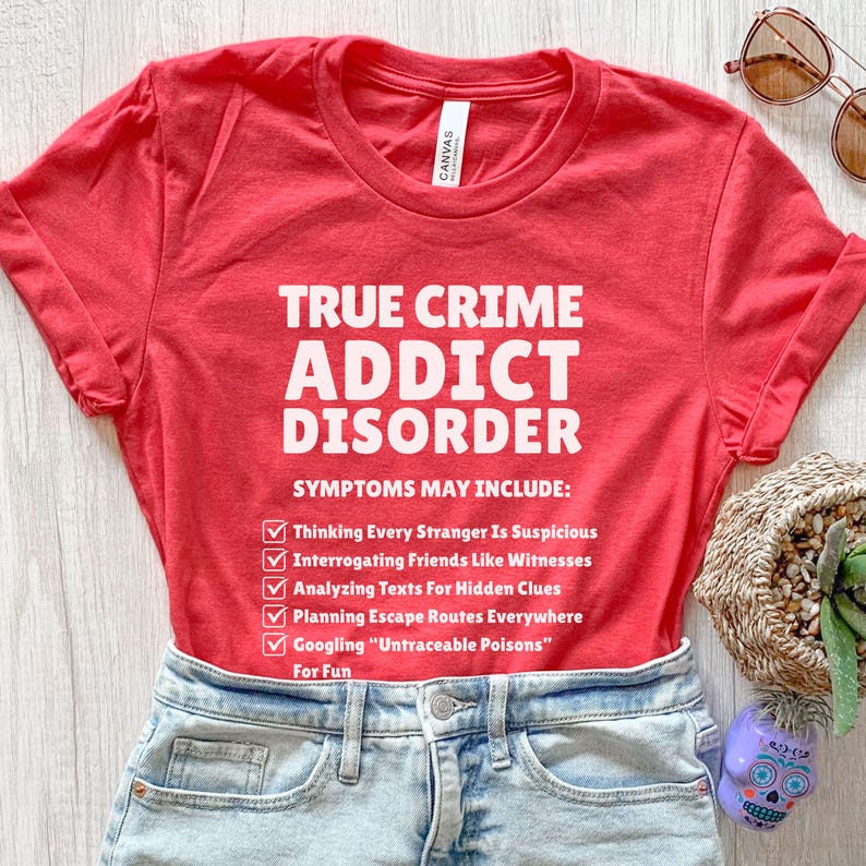 May include: A red t-shirt with the text "TRUE CRIME ADDICT DISORDER" in white. The shirt lists symptoms including "Thinking Every Stranger Is Suspicious" and "Googling 'Untraceable Poisons' For Fun". The shirt is paired with denim shorts.