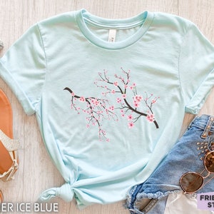 Puede incluir: Una camiseta azul claro con un gr&aacute;fico de una rama de cerezo en flor con flores rosas. La camiseta est&aacute; anudada en la parte inferior y est&aacute; sobre una superficie de madera.  HEATHER ICE BLUE est&aacute; escrito en la parte inferior de la imagen.