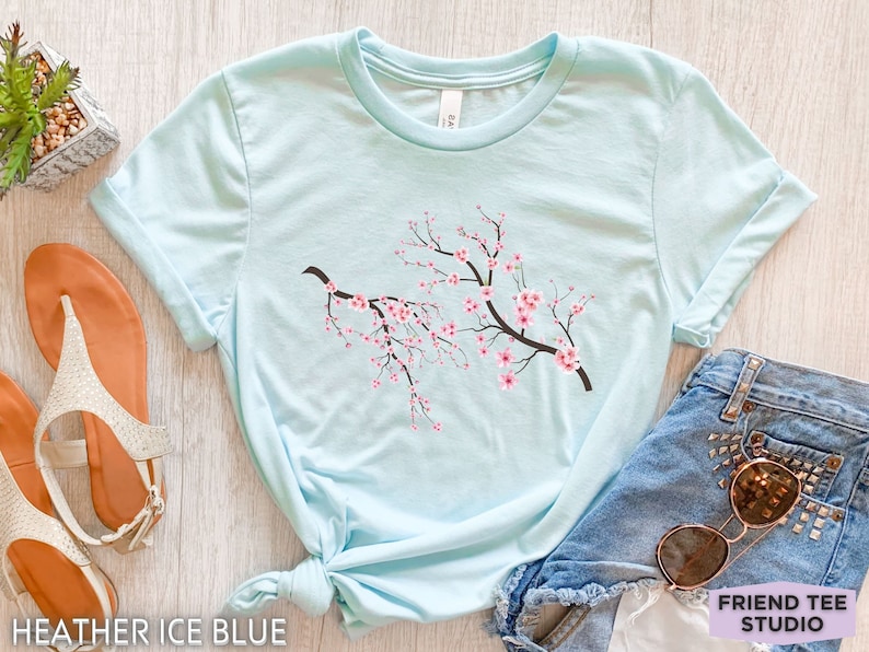 Puede incluir: Una camiseta azul claro con un gr&aacute;fico de una rama de cerezo en flor con flores rosas. La camiseta est&aacute; anudada en la parte inferior y est&aacute; sobre una superficie de madera.  HEATHER ICE BLUE est&aacute; escrito en la parte inferior de la imagen.