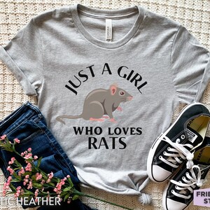 May include: An athletic heather gray short-sleeve t-shirt featuring a graphic of a brown rat centered between the black text that reads JUST A GIRL WHO LOVES RATS. The shirt is paired with dark blue denim jeans and black and white sneakers.