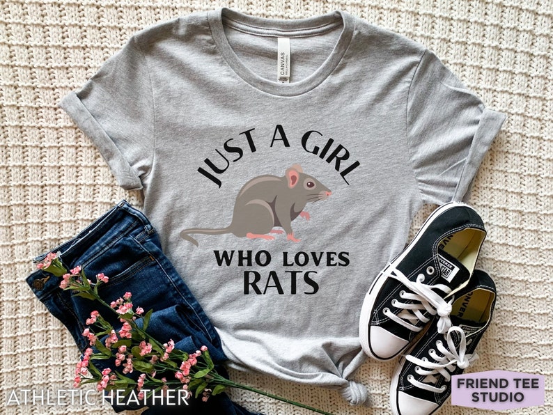 May include: An athletic heather gray short-sleeve t-shirt featuring a graphic of a brown rat centered between the black text that reads JUST A GIRL WHO LOVES RATS. The shirt is paired with dark blue denim jeans and black and white sneakers.