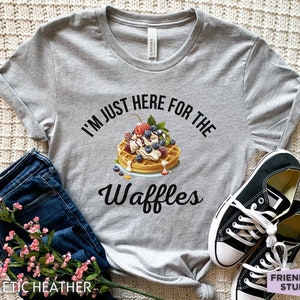 Op de afbeelding: Een grijs T-shirt met de tekst "I'm just here for the waffles" en een afbeelding van een wafel met slagroom, fruit en siroop.