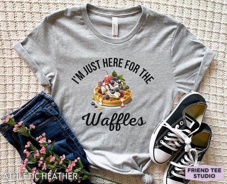 Op de afbeelding: Een grijs T-shirt met de tekst "I'm just here for the waffles" en een afbeelding van een wafel met slagroom, fruit en siroop.