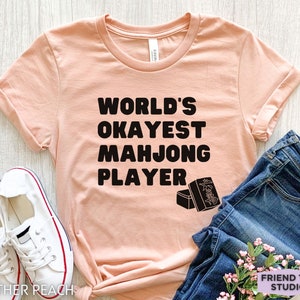 May include: A peach-colored t-shirt with black text that reads "World's Okayest Mahjong Player". The shirt has a graphic of mahjong tiles on the bottom right corner.
