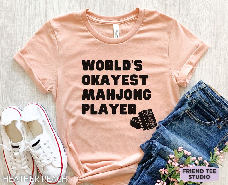 May include: A peach-colored t-shirt with black text that reads "World's Okayest Mahjong Player". The shirt has a graphic of mahjong tiles on the bottom right corner.