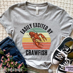 Op de afbeelding: Een grijs T-shirt met de tekst "Easily Excited By Crawfish" in zwarte letters. De tekst wordt omringd door een retro-zonnenondergangafbeelding met een rode kreeft in het midden.