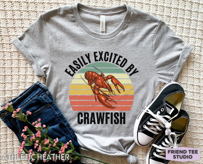 Op de afbeelding: Een grijs T-shirt met de tekst "Easily Excited By Crawfish" in zwarte letters. De tekst wordt omringd door een retro-zonnenondergangafbeelding met een rode kreeft in het midden.