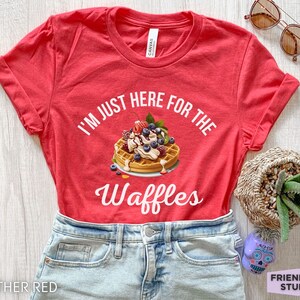 Op de afbeelding: Rood T-shirt met de tekst "I'm just here for the waffles" in wit. Een wafel met slagroom, blauwe bessen en aardbeien is afgebeeld.