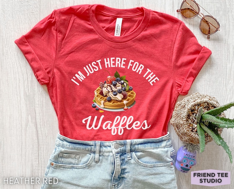 Op de afbeelding: Rood T-shirt met de tekst "I'm just here for the waffles" in wit. Een wafel met slagroom, blauwe bessen en aardbeien is afgebeeld.