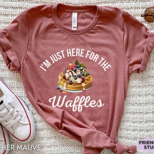 Op de afbeelding: Een heidekleurig T-shirt met de tekst "I'm just here for the waffles" in wit. Op het shirt is een wafel met slagroom, bessen en een kers afgebeeld.