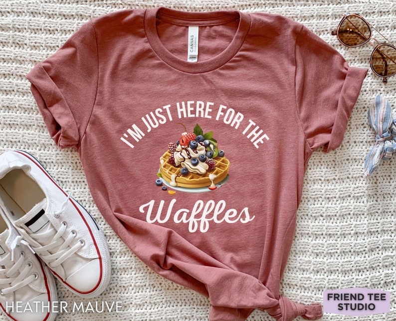 Op de afbeelding: Een heidekleurig T-shirt met de tekst "I'm just here for the waffles" in wit. Op het shirt is een wafel met slagroom, bessen en een kers afgebeeld.