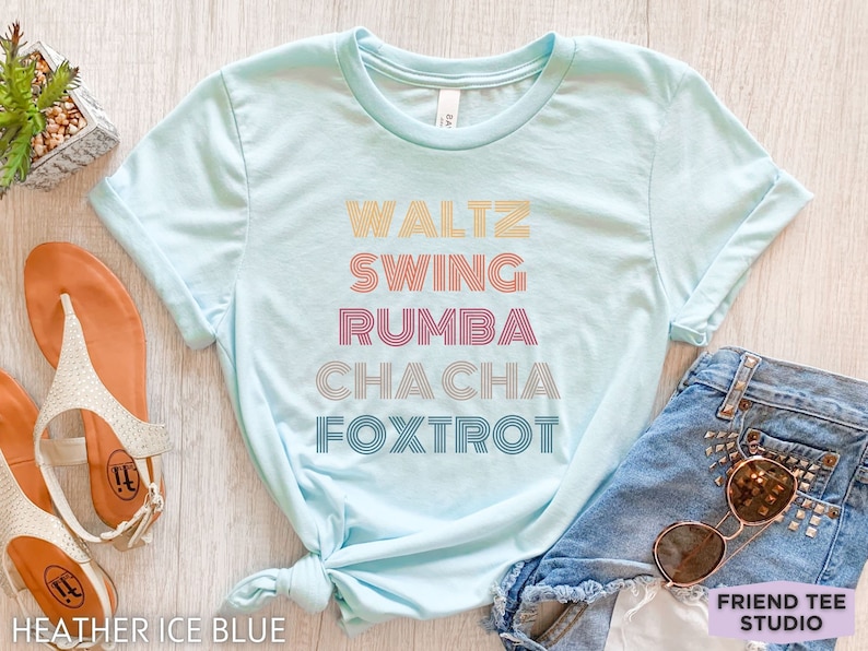 May include: A light blue t-shirt with the words "WALTZ, SWING, RUMBA, CHACHA, FOXTROT" in a retro font. The shirt is next to a pair of sandals and denim shorts. The text "HEATHER ICE BLUE" is at the bottom left.
