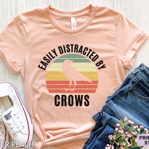 May include: A peach colored t-shirt with a retro sunset graphic and a black crow silhouette. The text "Easily Distracted By Crows" is printed on the shirt.