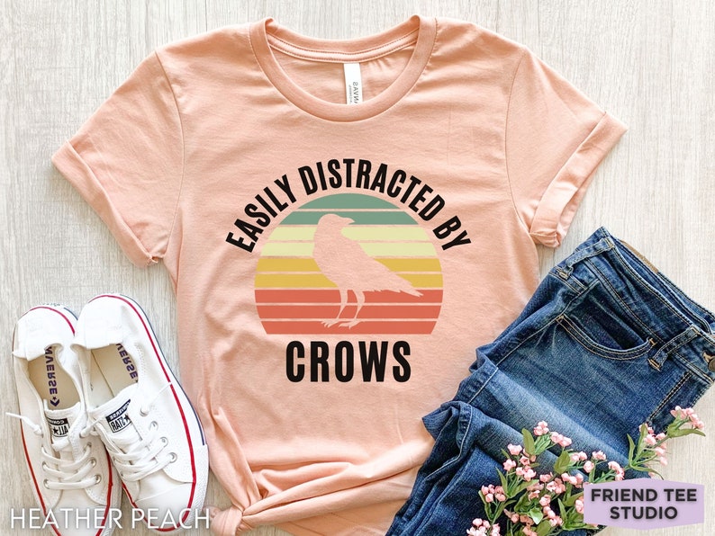May include: A peach colored t-shirt with a retro sunset graphic and a black crow silhouette. The text "Easily Distracted By Crows" is printed on the shirt.