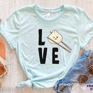 Op de afbeelding: Een lichtblauw T-shirt met het woord "LOVE" in zwarte letters. De "O" in LOVE is vervangen door een cartoon-dumpling met eetstokjes. De tekst "FRIEND TEE STUDIO" is zichtbaar in de rechterbenedenhoek.
