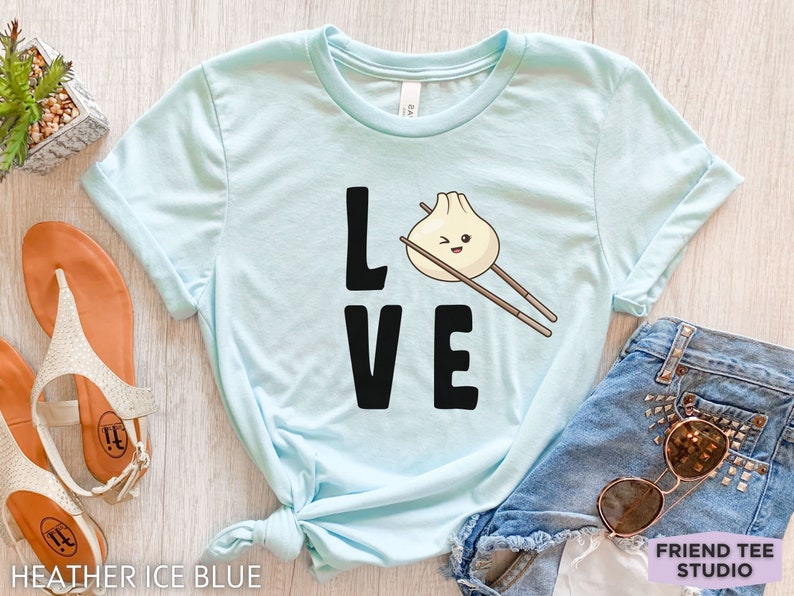 Op de afbeelding: Een lichtblauw T-shirt met het woord "LOVE" in zwarte letters. De "O" in LOVE is vervangen door een cartoon-dumpling met eetstokjes. De tekst "FRIEND TEE STUDIO" is zichtbaar in de rechterbenedenhoek.