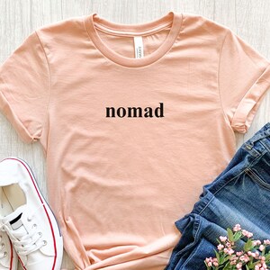 May include: A peach-coloured t-shirt with the word "nomad" printed in black. The shirt has folded sleeves and is paired with blue jeans and white trainers with red accents. A small bouquet of pink flowers is also visible.