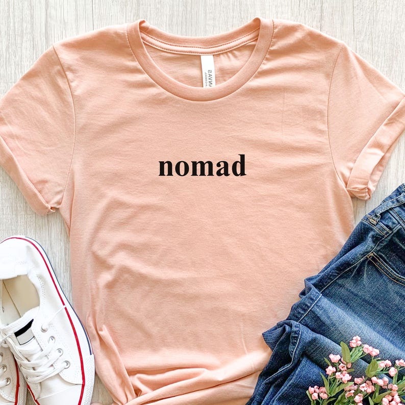 May include: A peach-coloured t-shirt with the word "nomad" printed in black. The shirt has folded sleeves and is paired with blue jeans and white trainers with red accents. A small bouquet of pink flowers is also visible.
