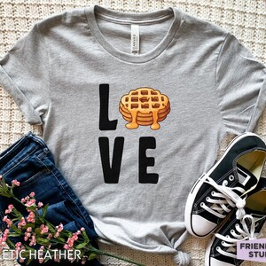 K&ouml;nnte beinhalten: Ein graues T-Shirt mit dem Wort "LOVE" in Schwarz bedruckt. Das "O" in LOVE wird durch ein Cartoon-Bild eines Waffels mit Sirup ersetzt.