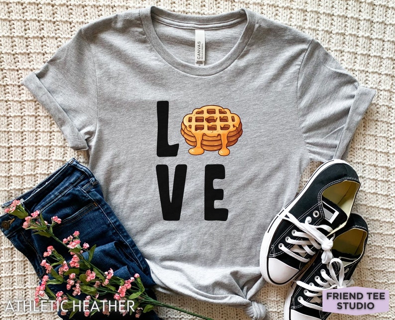 K&ouml;nnte beinhalten: Ein graues T-Shirt mit dem Wort "LOVE" in Schwarz bedruckt. Das "O" in LOVE wird durch ein Cartoon-Bild eines Waffels mit Sirup ersetzt.