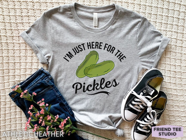 May include: Athletic heather gray t-shirt with the text "I'M JUST HERE FOR THE PICKLES" and a graphic of two green pickles. The shirt is paired with blue jeans and black sneakers.