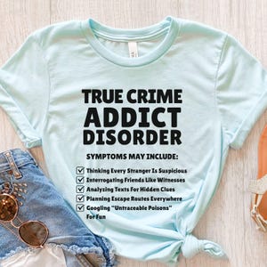May include: Pale blue t-shirt featuring the text "TRUE CRIME ADDICT DISORDER" in black. The shirt lists symptoms such as suspecting strangers, interrogating friends, and Googling "Untraceable Poisons".