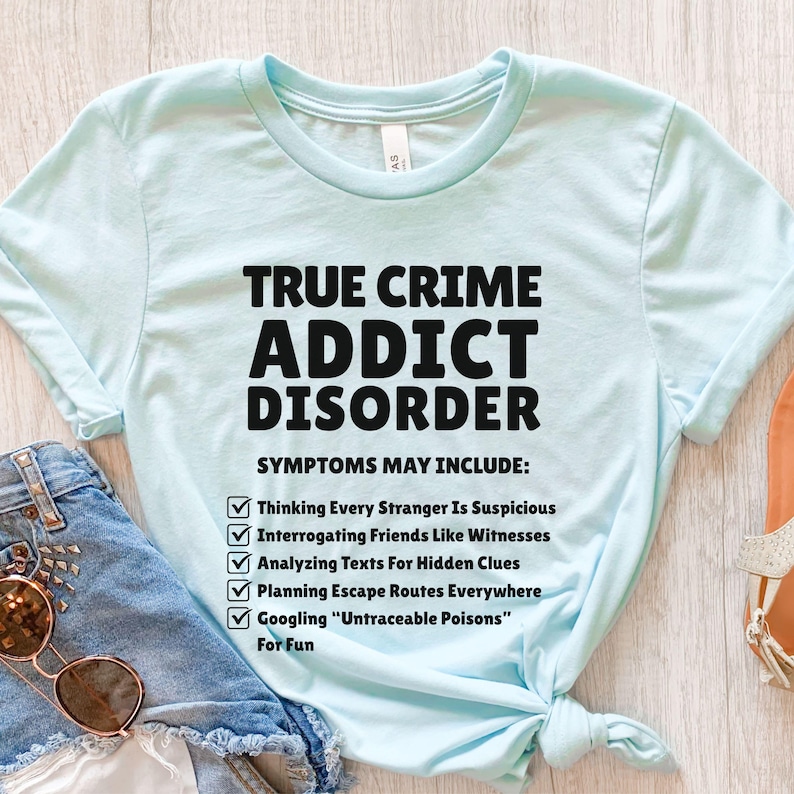May include: Pale blue t-shirt featuring the text "TRUE CRIME ADDICT DISORDER" in black. The shirt lists symptoms such as suspecting strangers, interrogating friends, and Googling "Untraceable Poisons".