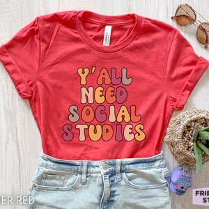 May include: A red t-shirt with the text "Y'all Need Social Studies" in a retro font with orange, pink, and brown colors.