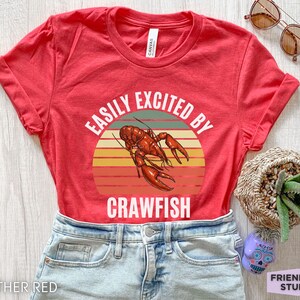Op de afbeelding: Rood T-shirt met een vintage-stijl grafische afbeelding van een rivierkreeft en de tekst "Easily Excited By Crawfish".