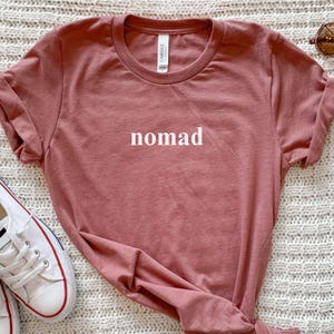 May include: A dusky pink t-shirt with the word "nomad" printed in white. The shirt has a crew neck and short sleeves. White canvas trainers with red and blue detailing are visible in the lower left corner.