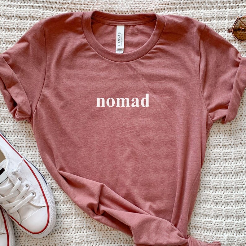 May include: A dusky pink t-shirt with the word "nomad" printed in white. The shirt has a crew neck and short sleeves. White canvas trainers with red and blue detailing are visible in the lower left corner.