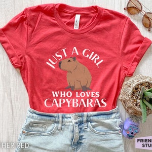 May include: Red t-shirt with white text that reads "Just a girl who loves capybaras". A cartoon capybara is pictured above the text.